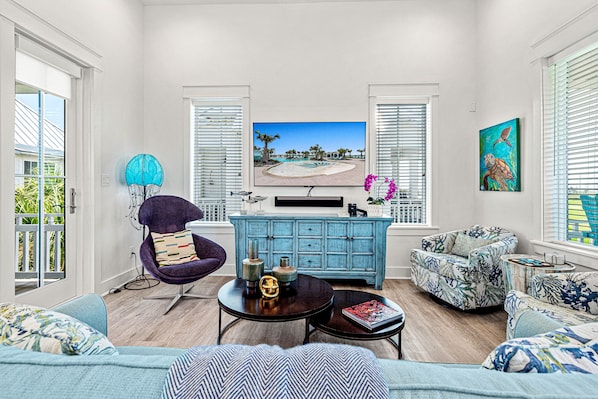 A living room with a large flat-screen TV, a blue cabinet, a purple chair, a sofa, and a coffee table. - Modern home w/ two master suites, golf course view - pool, beach, gym, & more (Port Aransas)
