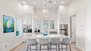 A modern kitchen with white cabinetry, a central island with bar stools, stainless steel appliances, and pendant lighting. - Modern home w/ two master suites, golf course view - pool, beach, gym, & more (Port Aransas)