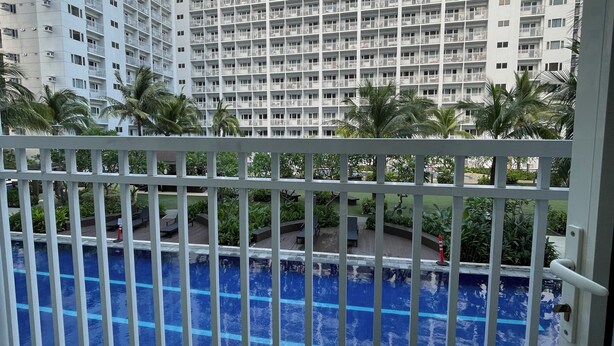 A view of a swimming pool with a white fence in the foreground, surrounded by greenery and palm trees. In the background, there is a tall residential building with multiple balconies.