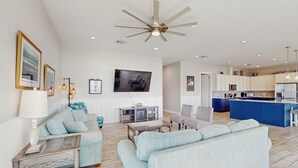 A spacious living room with a large sofa, a coffee table, and a TV mounted on the wall. There is a kitchen area with a blue island and white cabinets. - Stunning bay front home with fishing pier, large deck, & private pool (Rockport)