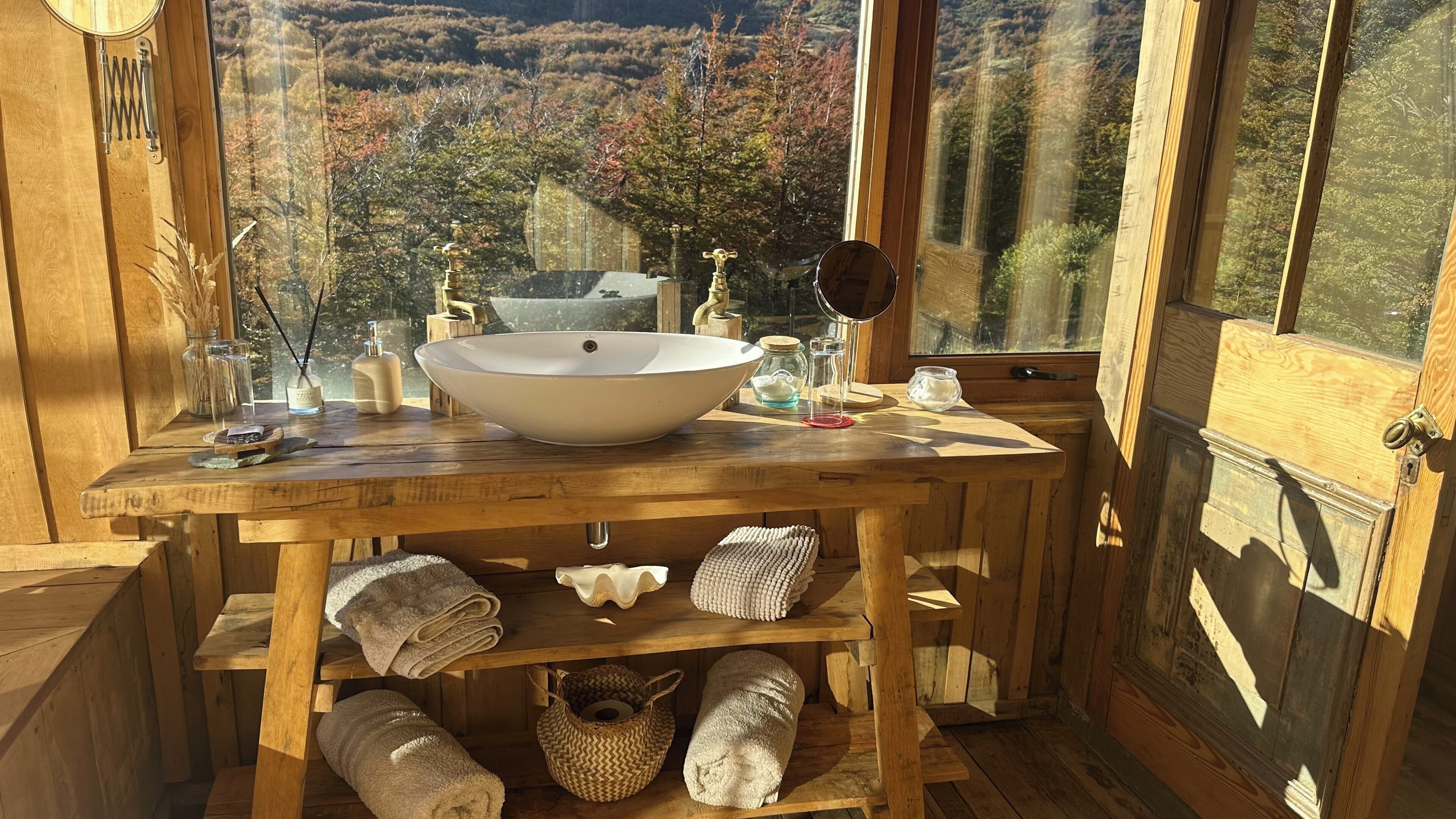 A wooden bathroom with a large sink, towels, and a view of autumn foliage.