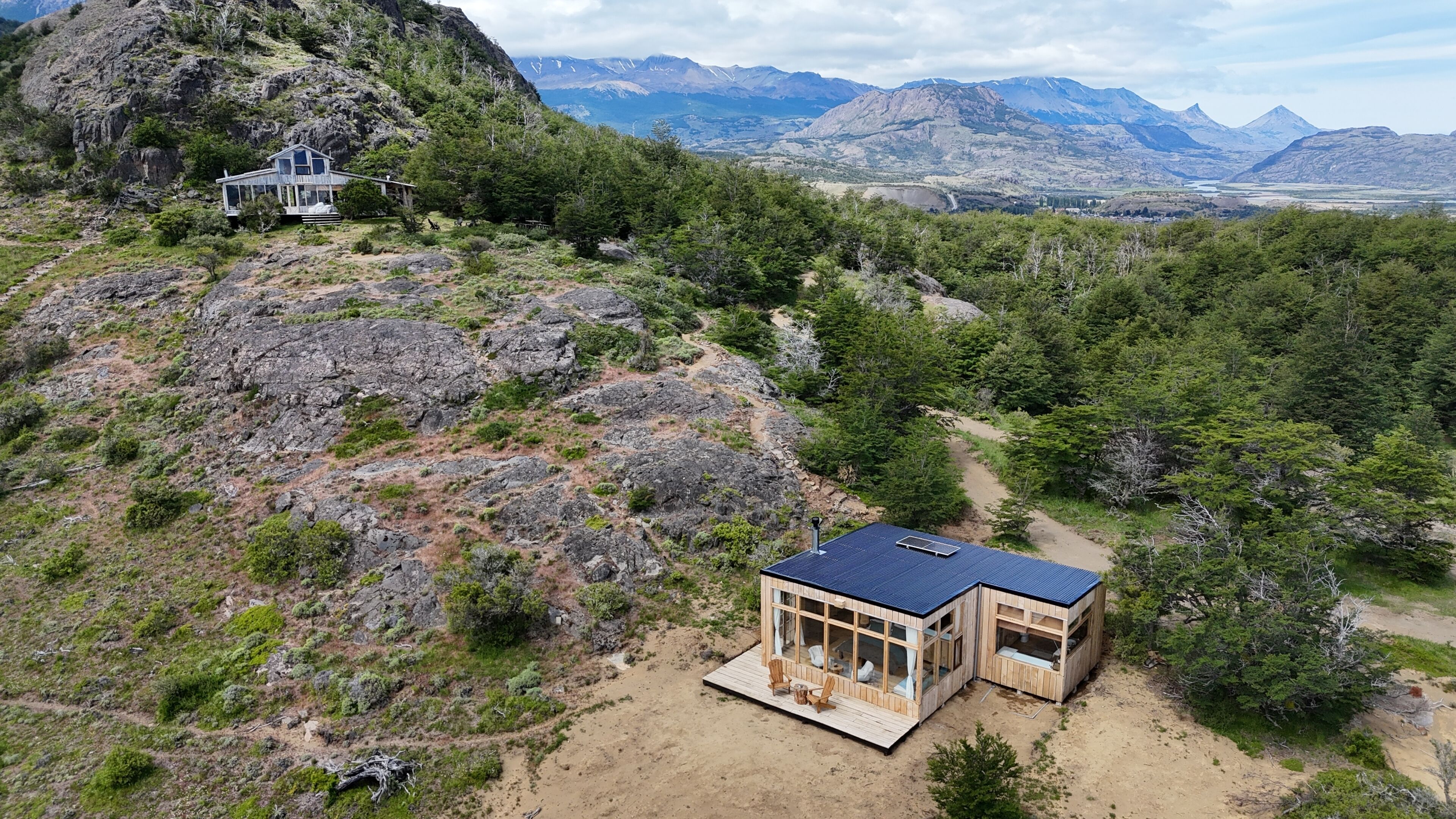 A modern wooden cabin with large windows, surrounded by rocky terrain and dense forest, with mountains in the background.