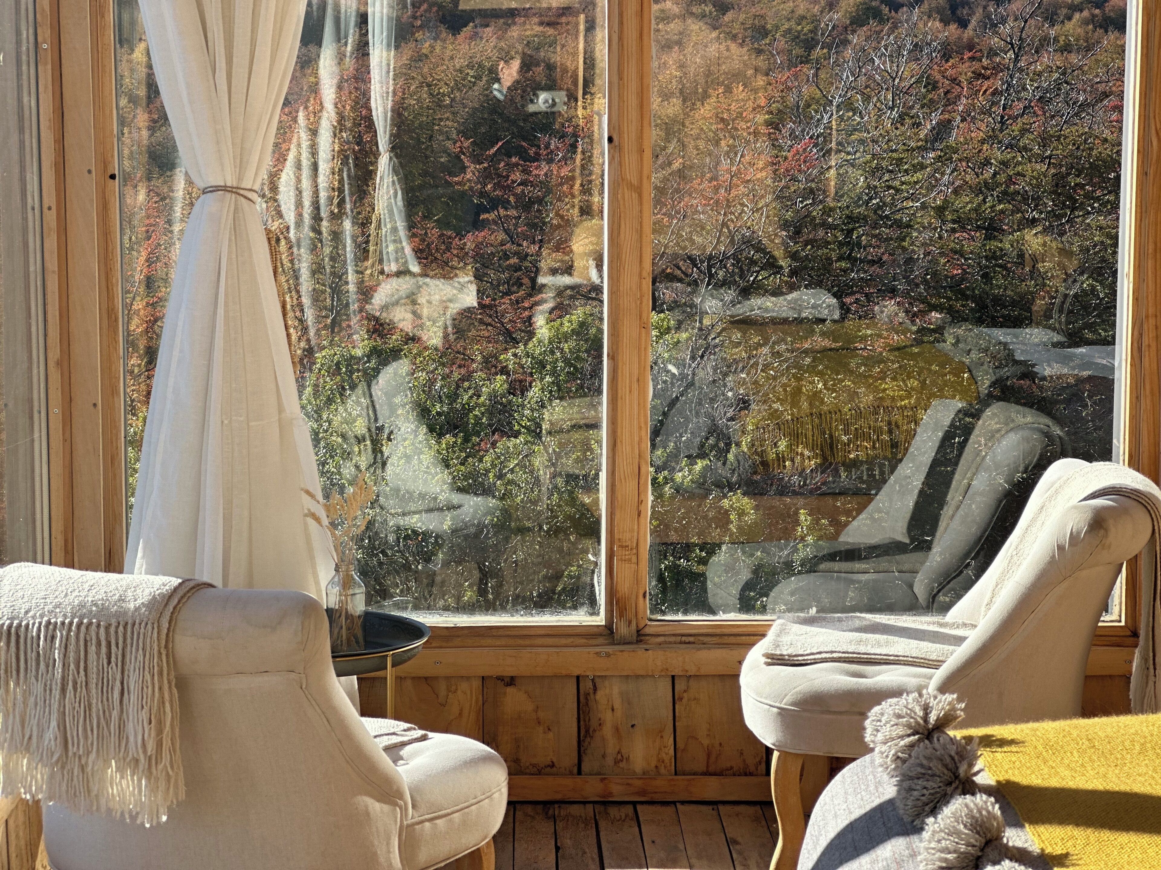Quarto com uma grande janela com vista para uma paisagem natural, duas cadeiras, uma mesa pequena e piso de madeira.