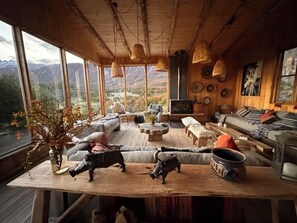 A cozy cabin interior with a large window offering a mountain view, wooden furniture, and decorative items.