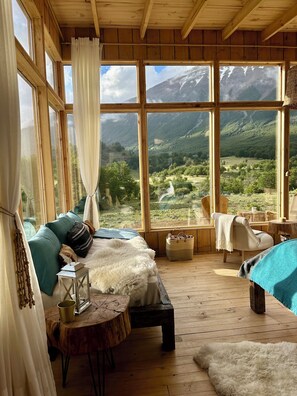 A cozy room with a bed, a small table, and a view of a mountainous landscape through large windows.