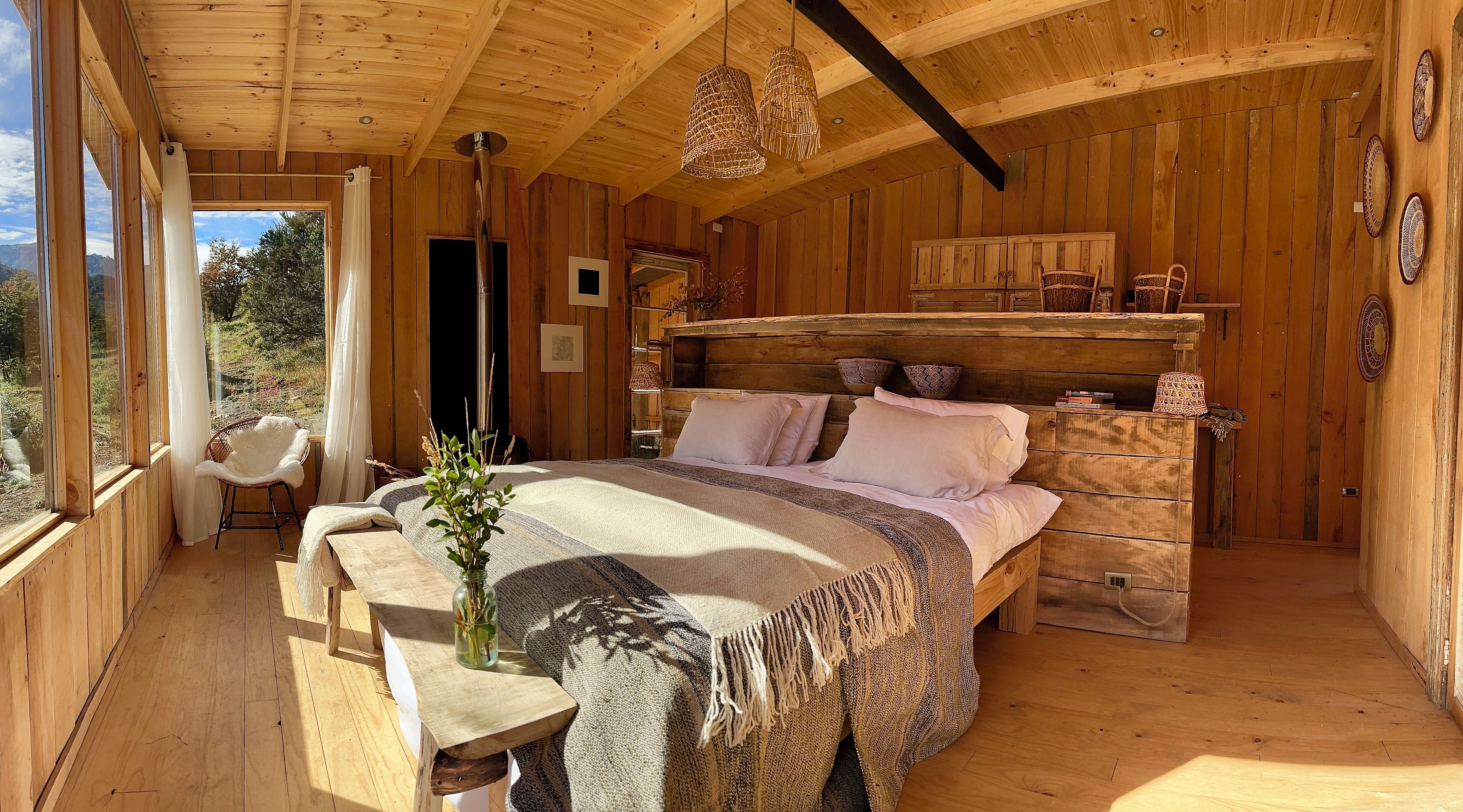 Interno di una baita in legno con un letto, una panca, una finestra vista e un lampadario.