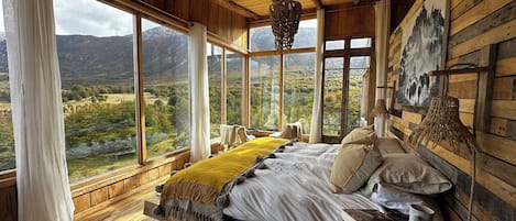 Une chambre à coucher rustique avec un grand lit, des murs en bois et une vue sur un paysage montagneux, visible à travers de grandes fenêtres.