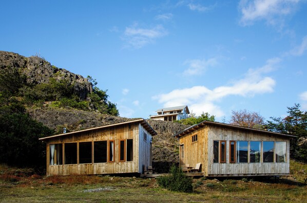 Two wooden cabins with large windows, set on a hillside with a clear sky above.
