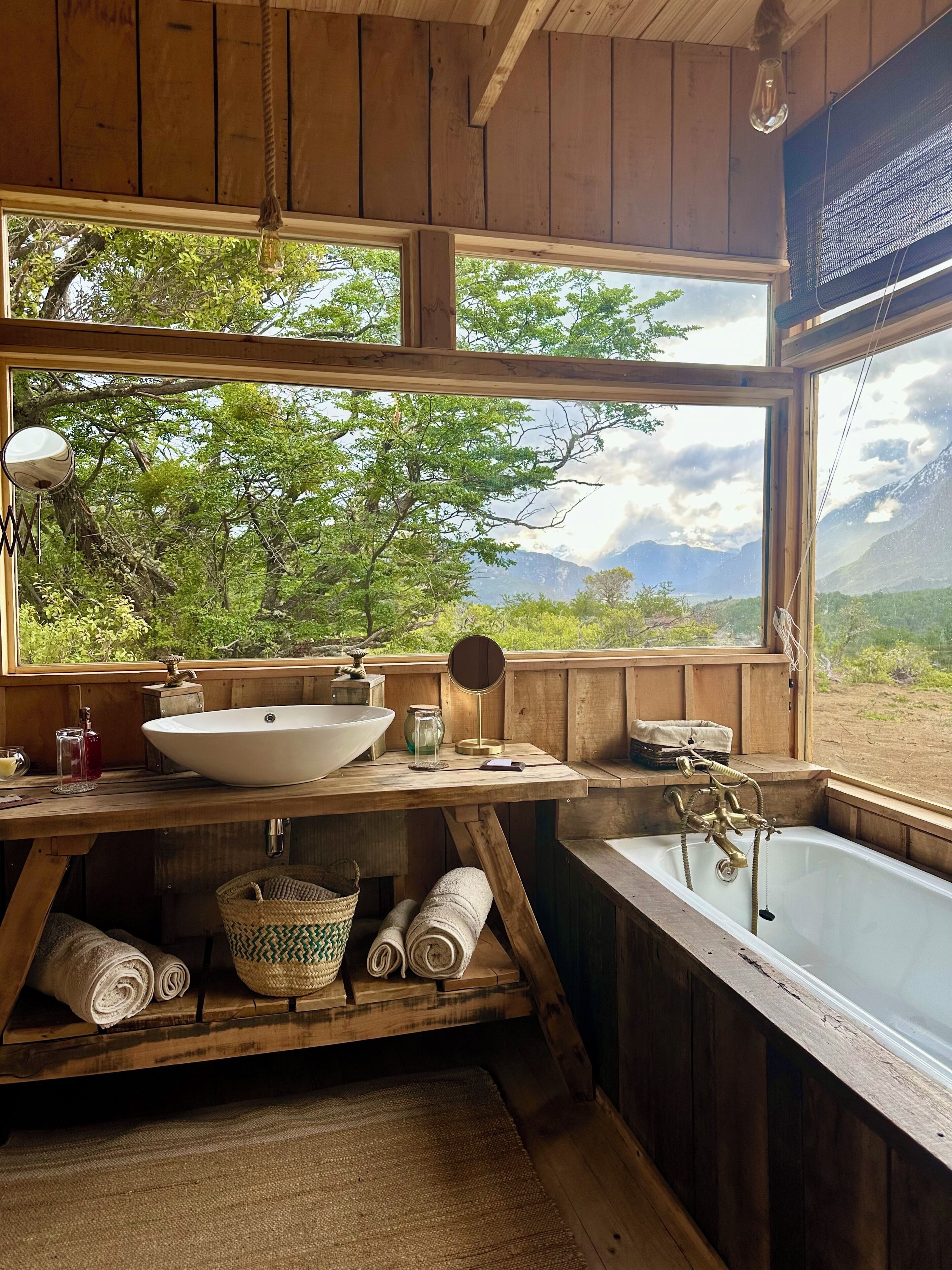 A rustic bathroom with a wooden interior, a large window offering a view of greenery and mountains, a sink, and a bathtub.