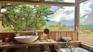 A rustic bathroom with a wooden interior, a large window offering a view of greenery and mountains, a sink, and a bathtub.
