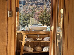 A wooden bathroom with a large sink, shelves with towels, and a view of autumn trees.