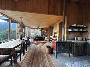 A wooden interior with a dining area, kitchen, and large windows overlooking a mountainous landscape.