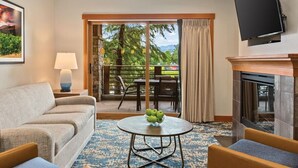 A hotel room with a sofa, a round coffee table with apples, a fireplace, a TV, and a balcony with outdoor seating.