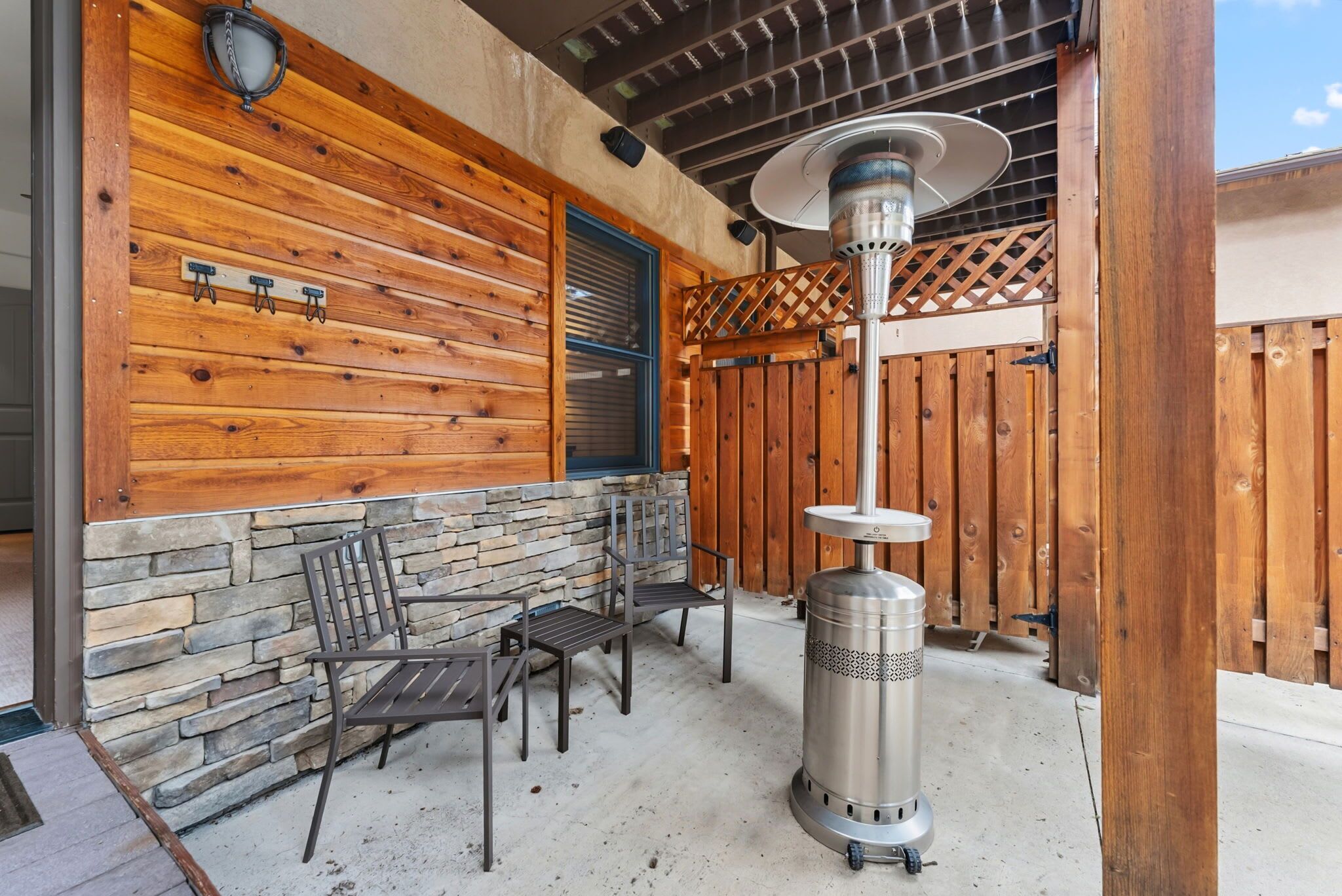 Een terras buiten met stenen en houten wanden, een metalen terrasverwarmer en twee metalen stoelen.