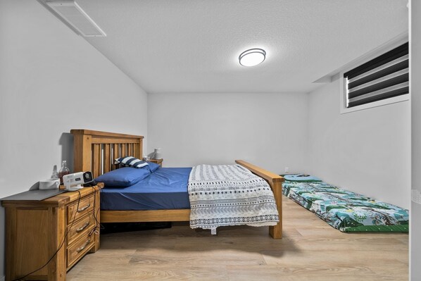 A bedroom with a wooden bed, a bedside table, a desk, and a window with blinds.