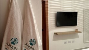 A hotel room with two towels hanging on a wall, a flat-screen TV mounted above a wooden shelf, and a bed with a green blanket. - Ámari, Spacious beach front 3-bedroom apartment with pool in sunny Puerto Plata (Puerto Plata)
