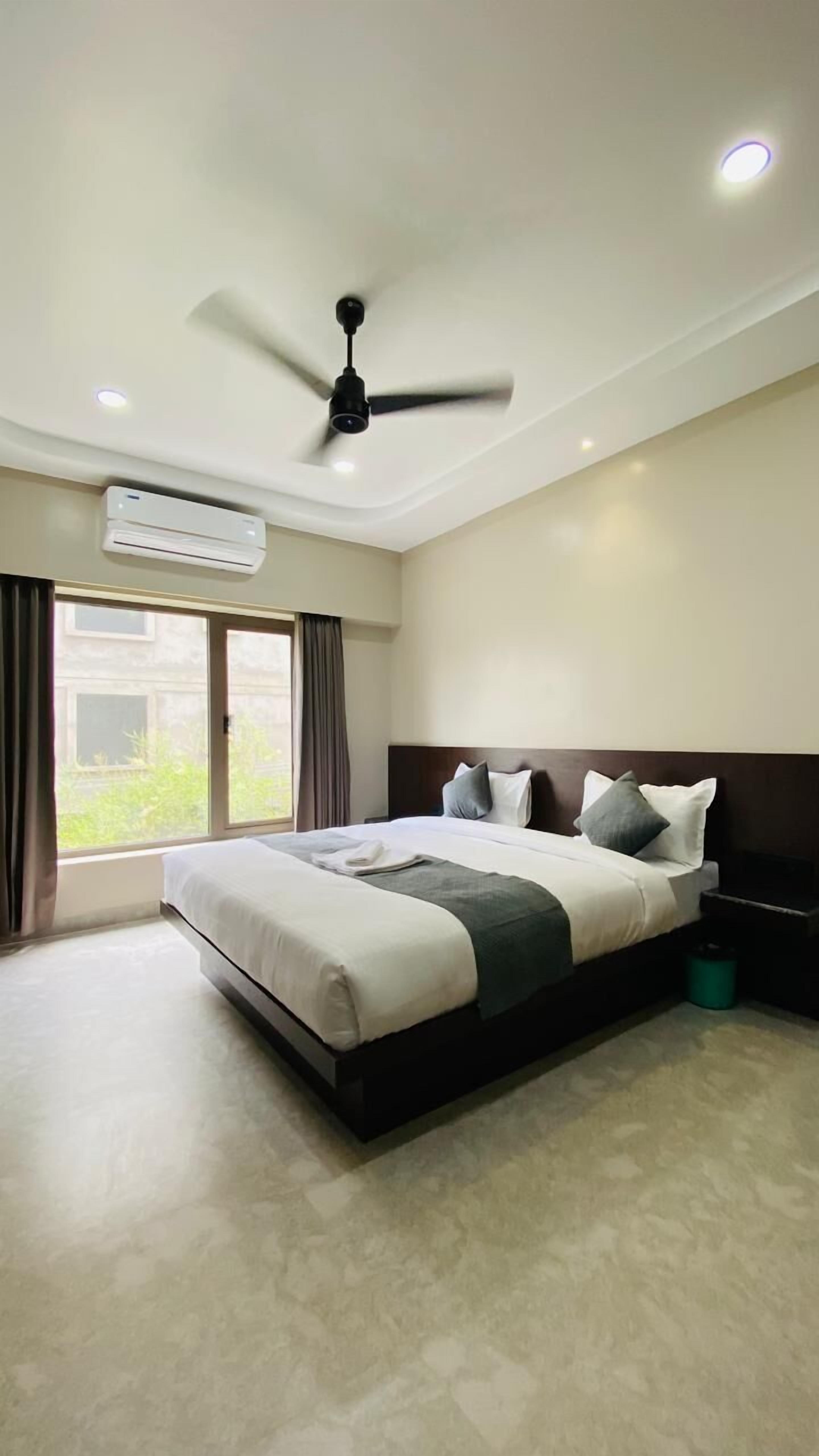 A modern hotel room with a large bed, ceiling fan, air conditioning unit, and a view of greenery through the window.