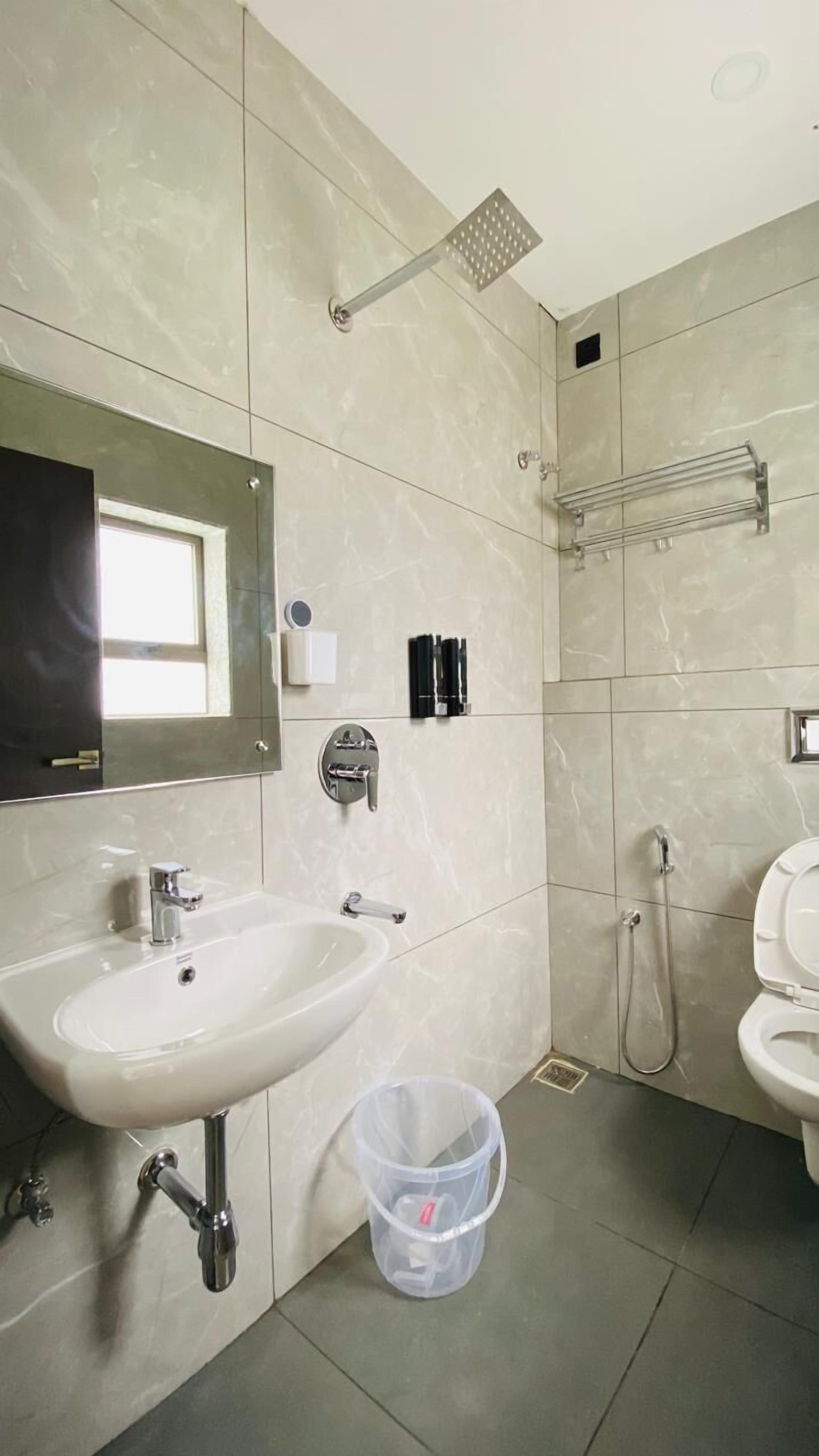 A modern bathroom with a white sink, mirror, and toilet. There is a shower area with a handheld showerhead and a towel rack. A trash bin is visible on the floor.
