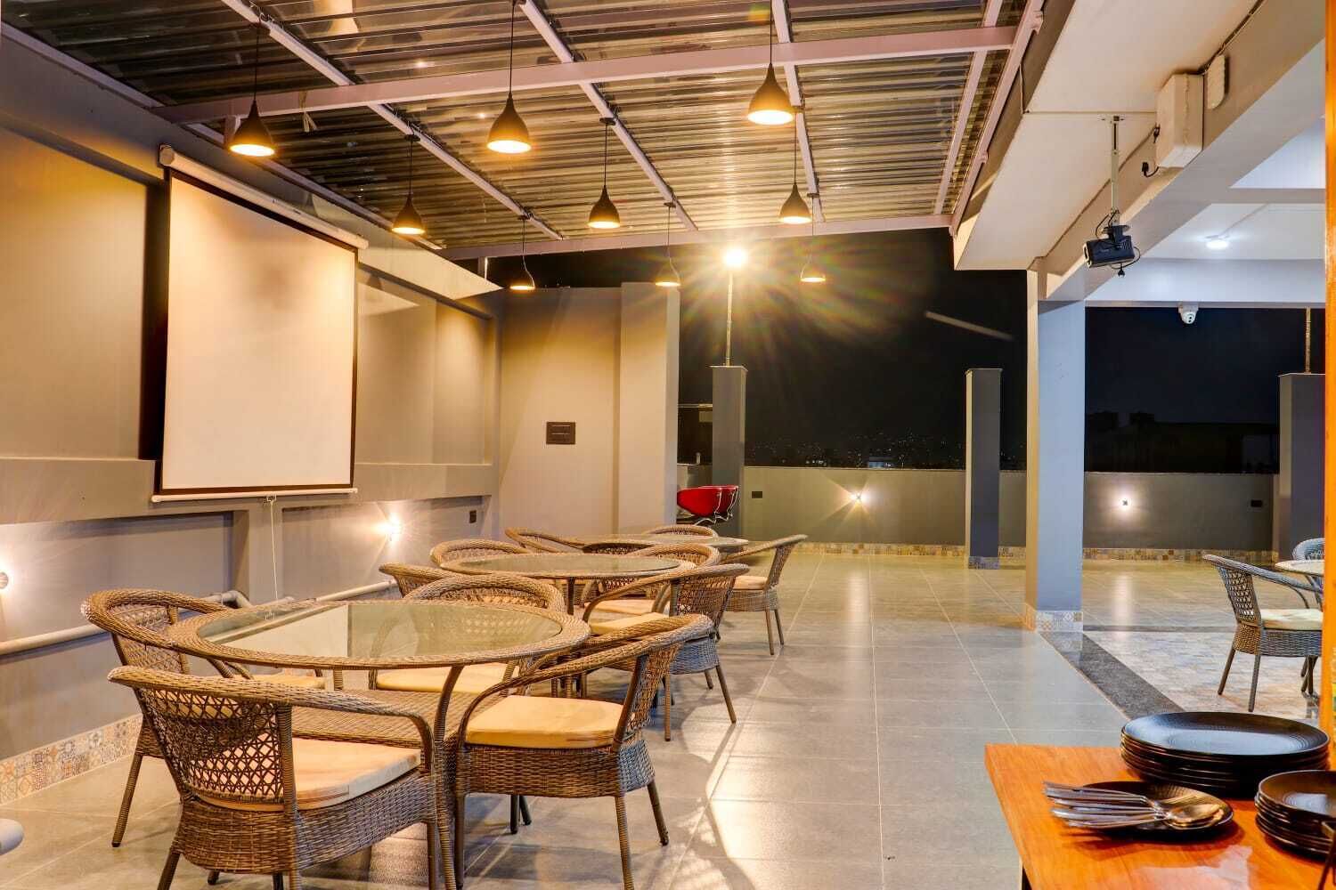 A rooftop dining area with wicker furniture, a projector screen, and a view of the city at night.