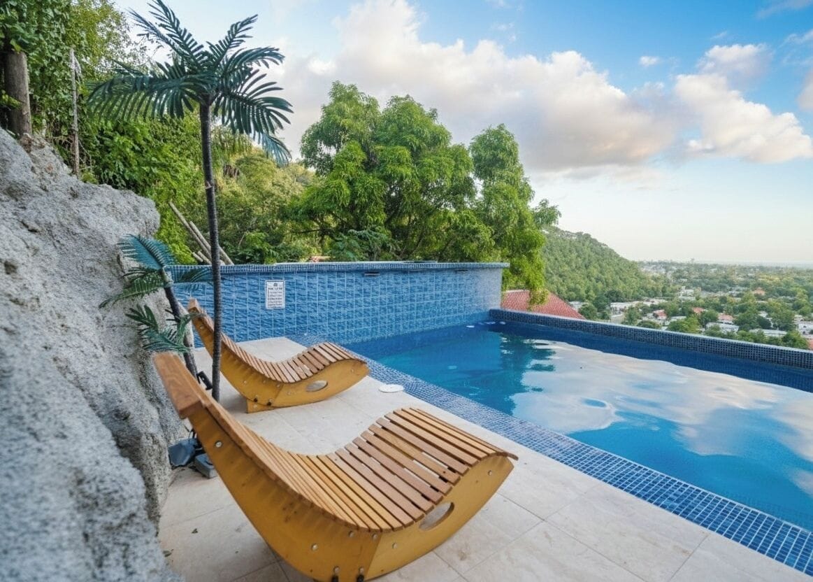 A swimming pool with wooden loungers, surrounded by a stone wall and lush greenery.