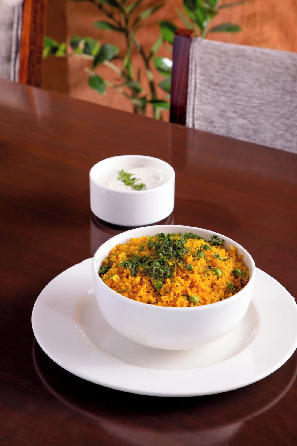 A bowl of yellow rice garnished with herbs on a white plate, with a small bowl of white sauce in the background.