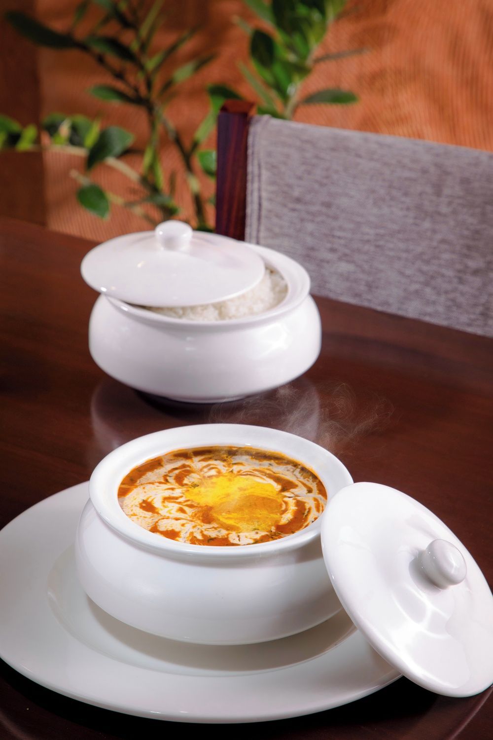 A white ceramic bowl with a lid on a white plate, placed on a wooden table.