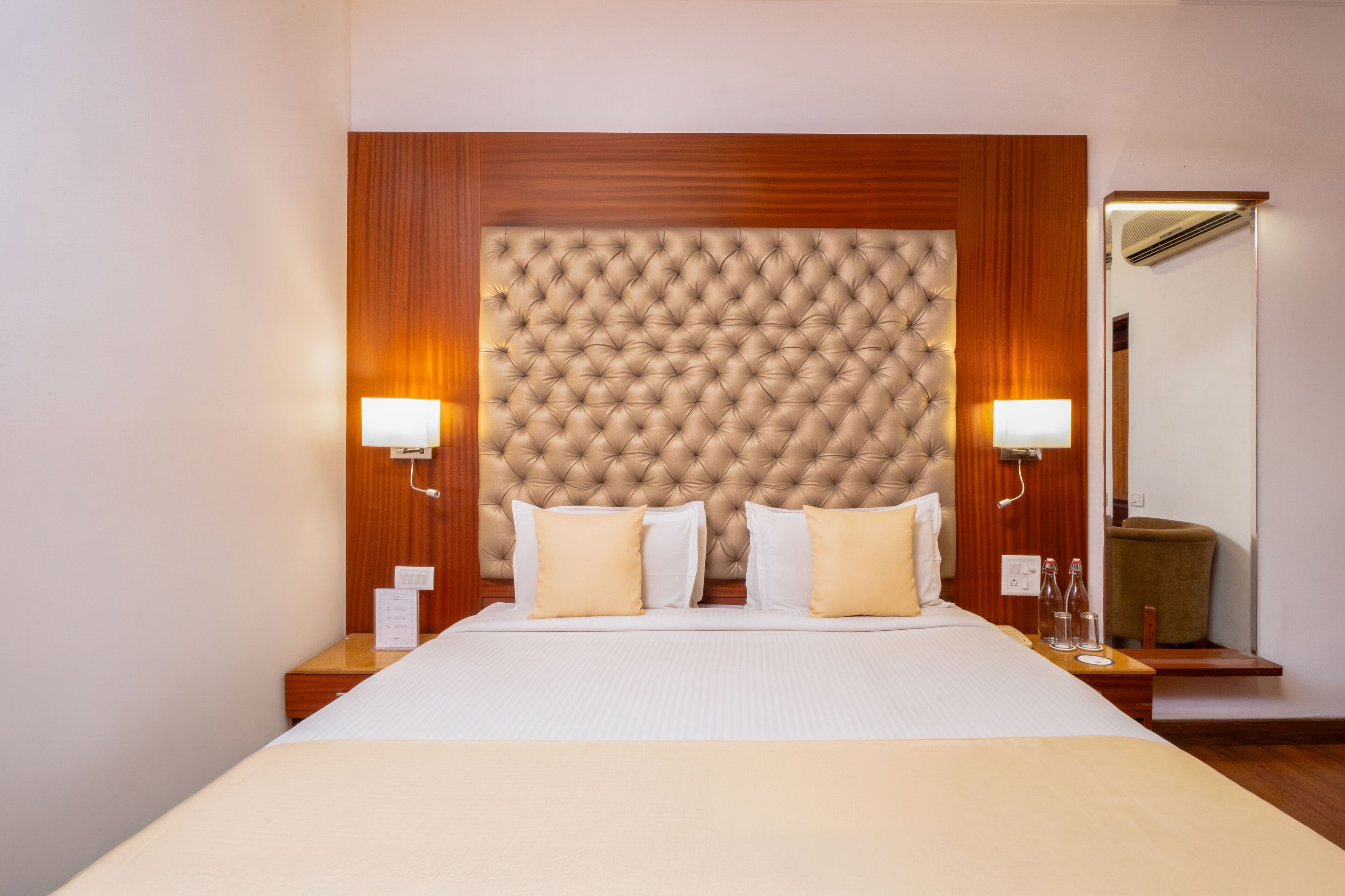 A hotel room with a large bed, wooden headboard, two bedside lamps, a nightstand, a mirror, and a small table with a bottle and glass.