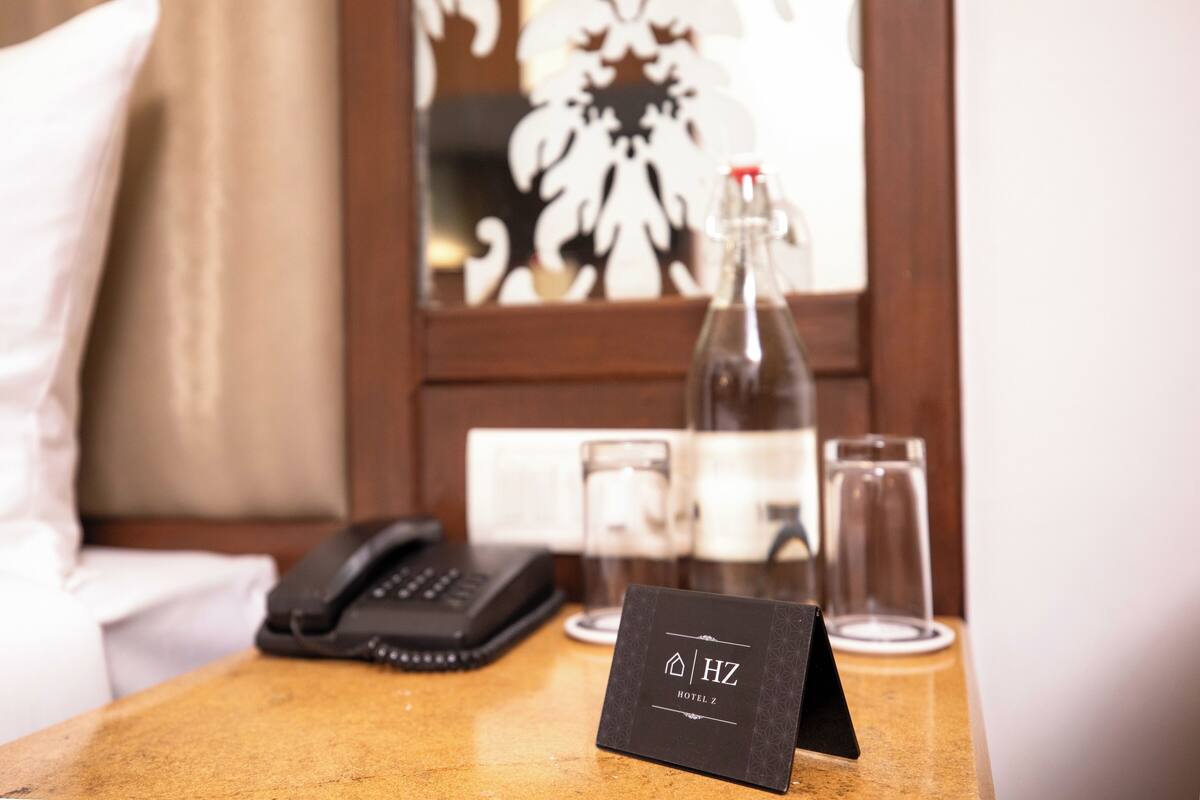 Une chambre d’hôtel avec un lit, un téléphone sur la table de chevet, un verre d’eau et une bouteille d’eau sur une table de chevet en bois.