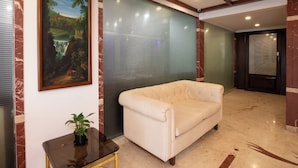 A lobby with a beige sofa, a framed landscape painting, a small table with a plant, and a glass partition with horizontal lines.