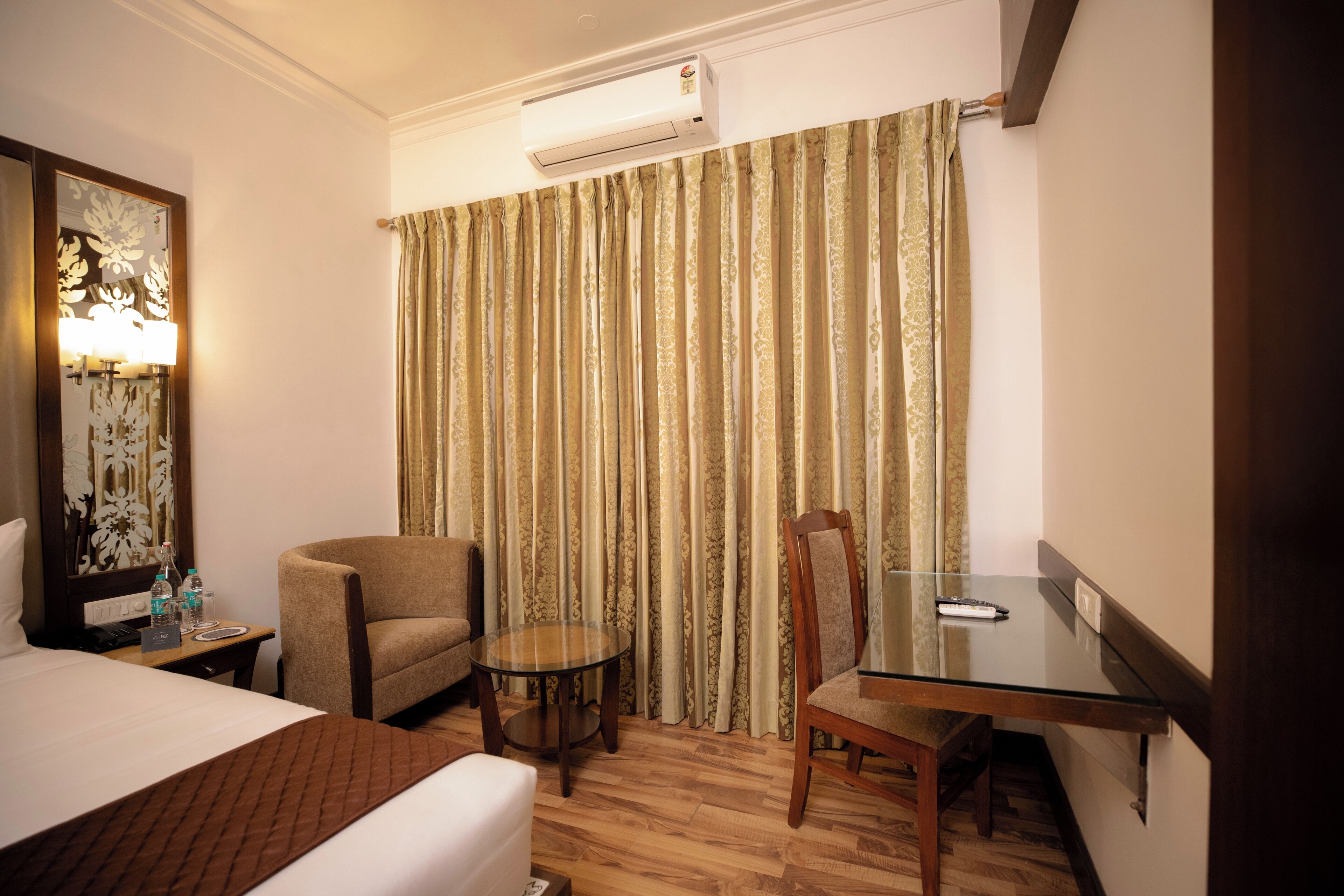 Una habitación de hotel con cama, una silla, una mesita redonda pequeña y una mesita de cristal, además de una cortina estampada.