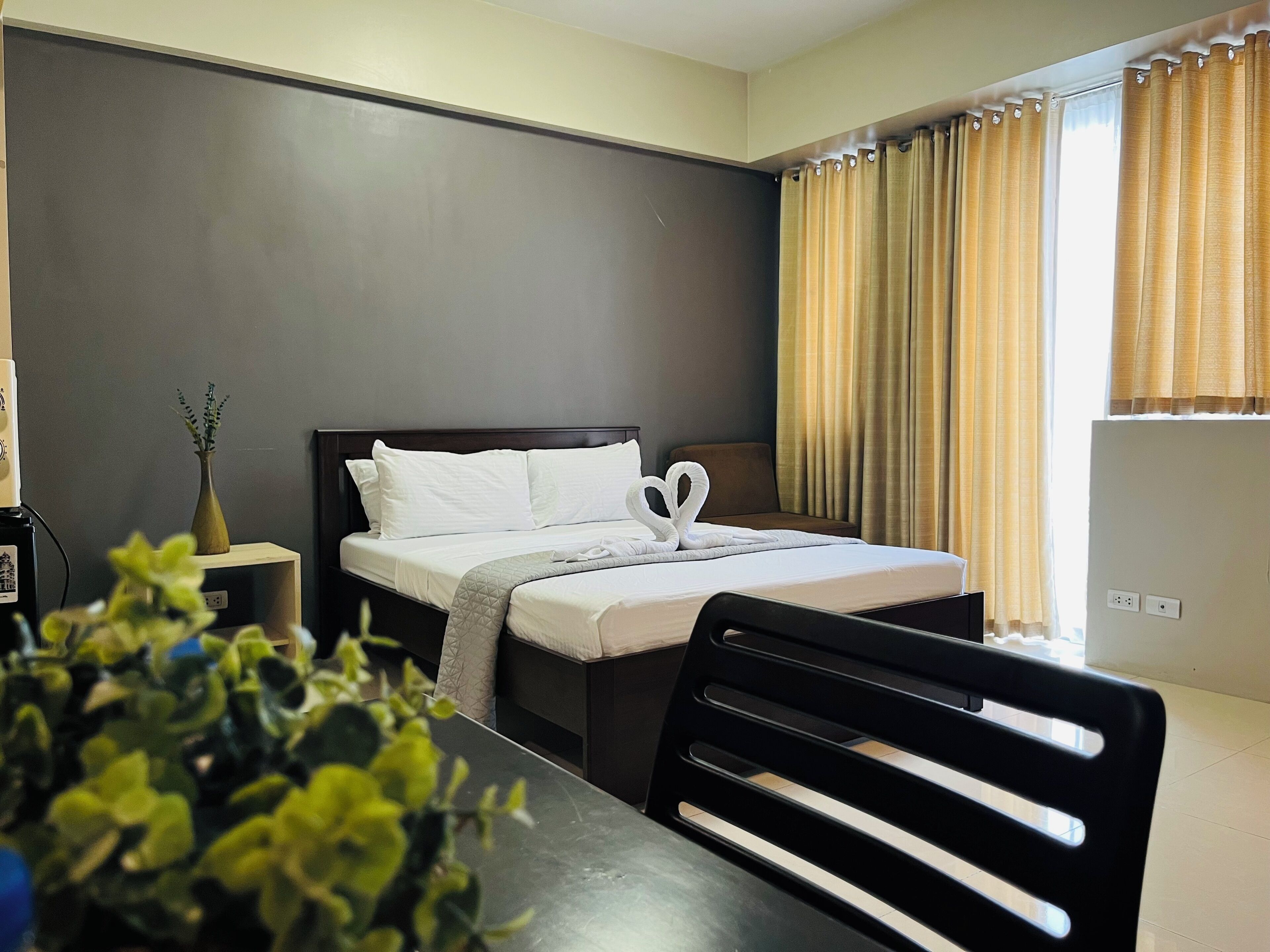 A hotel room with a bed, a bedside table, a chair, a potted plant, and a window with curtains.