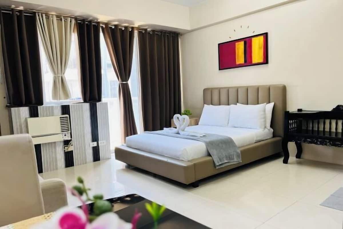 A hotel room with a bed, a desk, a chair, a window with curtains, an air conditioning unit, and a wall-mounted artwork.