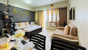 A hotel room with a bed, a desk, a chair, a TV, a ceiling fan, and a window with curtains. - One Palm Tree Villas (Pasay)