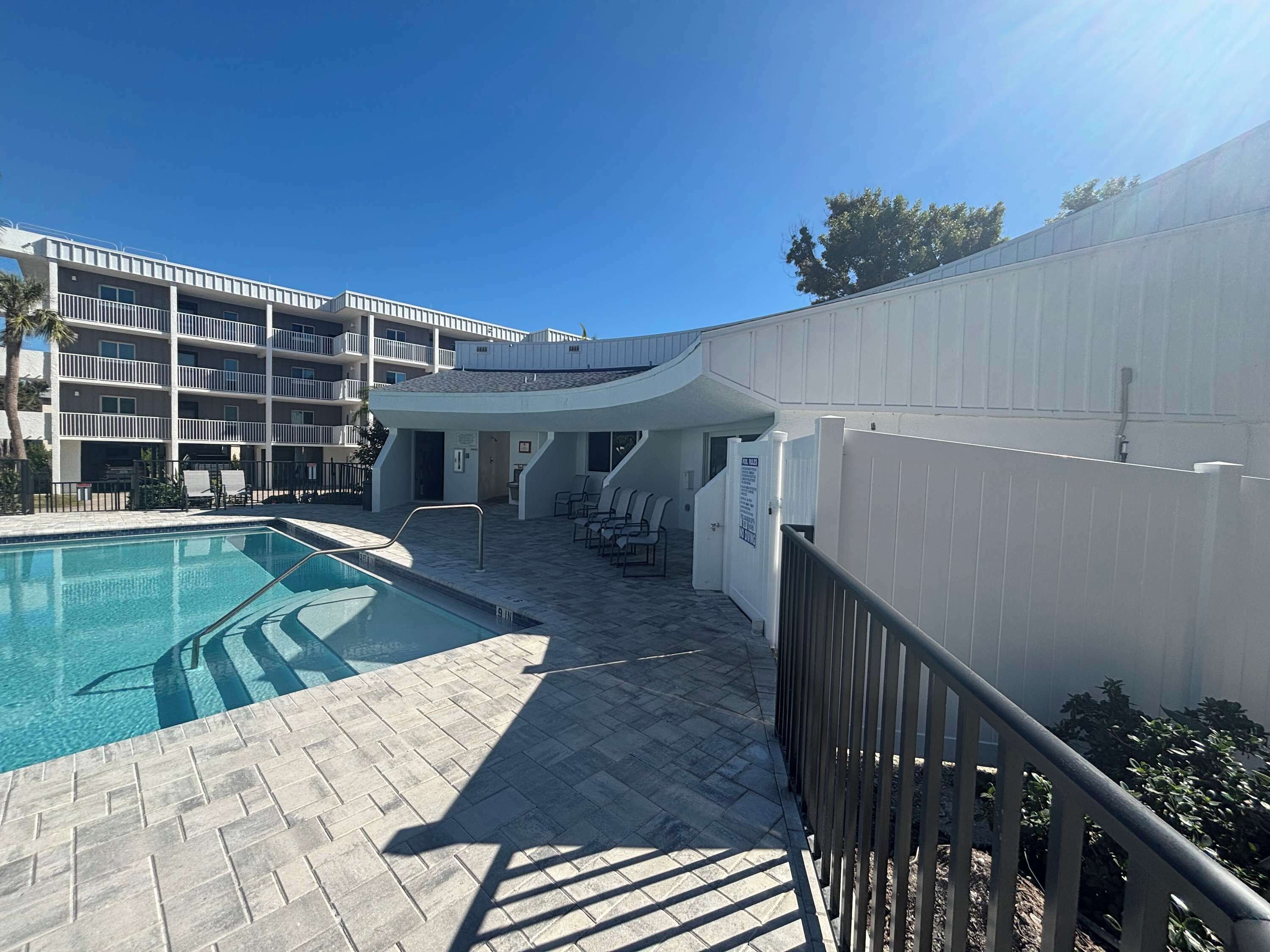 A swimming pool with a tiled deck, a white building with balconies, and a clear blue sky.