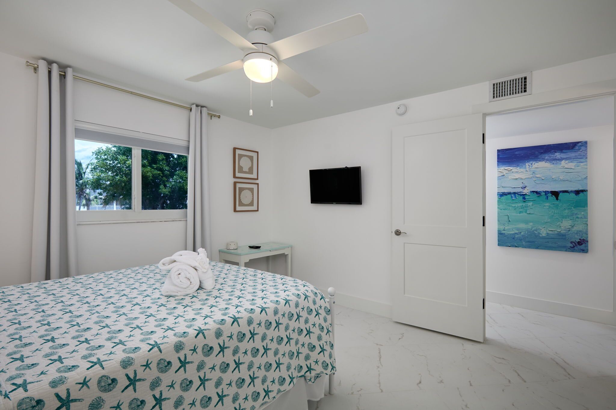 Une chambre d’hôtel avec un lit, un ventilateur de plafond, une télévision, un tableau et une fenêtre avec des rideaux.