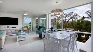 A modern living room with a TV, seating, and a dining area with a view of the outdoors. - Island Beach Club 340B | Spacious and Upscale Beach Residence | Pet-Friendly (Sanibel)