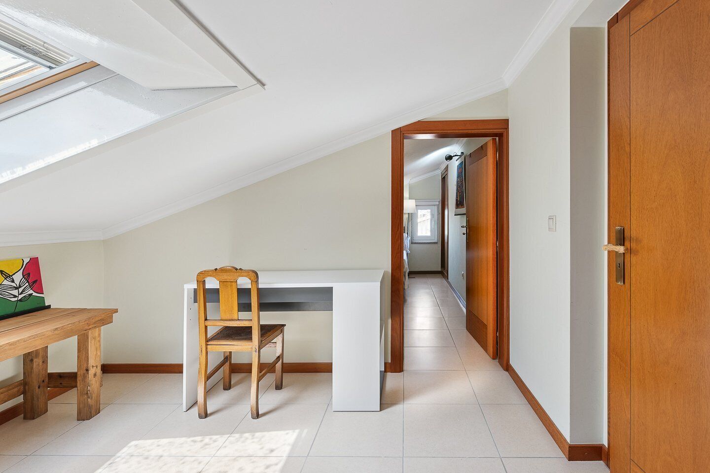 A room with a sloped ceiling, a wooden dining table, a chair, and a white desk.