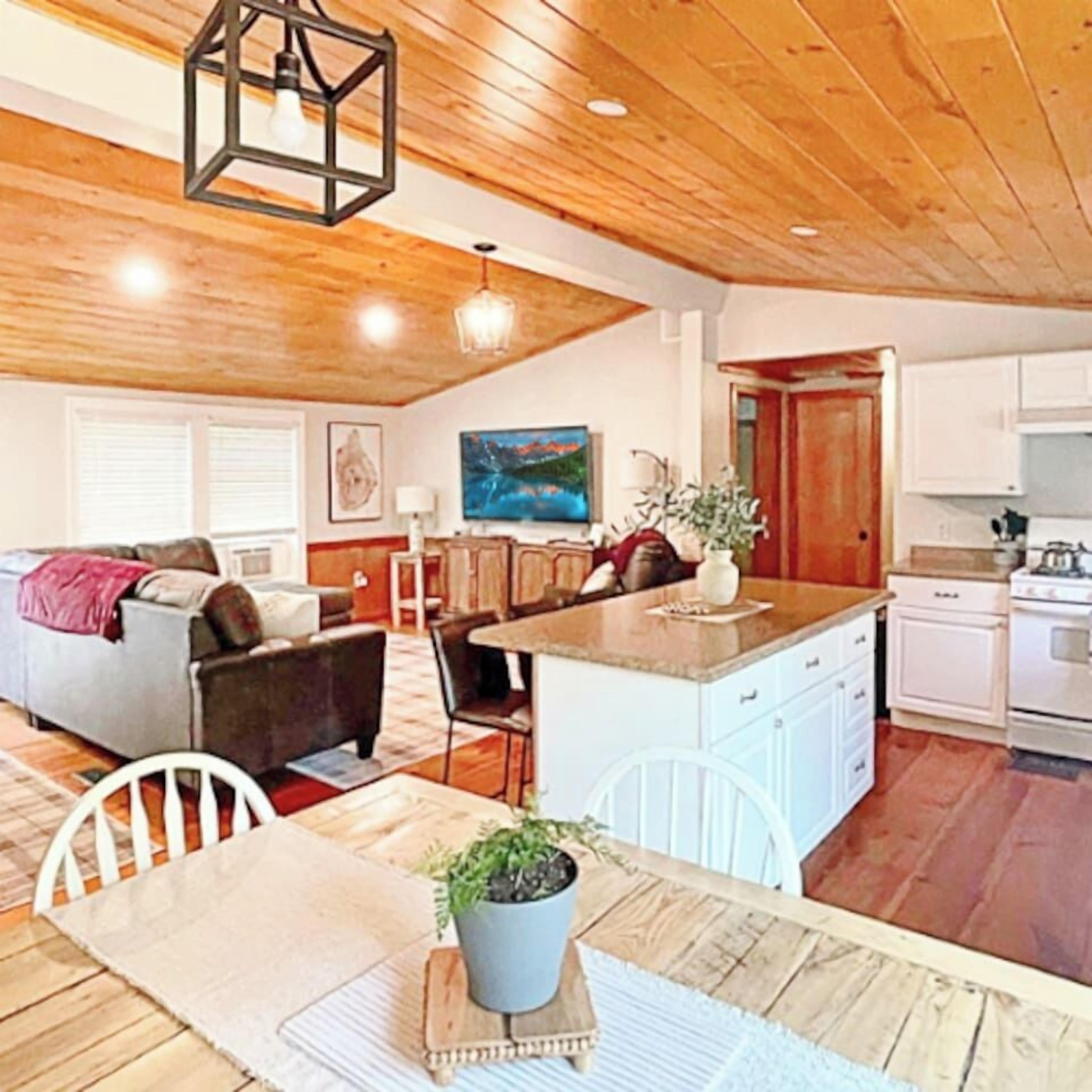 A cozy living room with a sofa, a dining table with chairs, a kitchen area with cabinets and appliances, and a wooden ceiling.