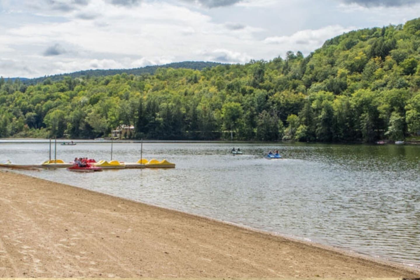A lakeside view with a sandy beach, a row of yellow floating docks, and a forested hill in the background.