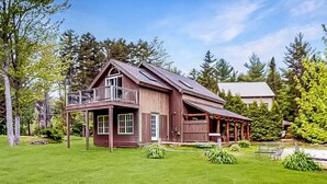Une maison en bois à deux étages, dotée d’un balcon, entourée d’une pelouse bien entretenue et d’arbres.
