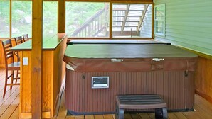 A hot tub area with a wooden deck, a table with chairs, and a view of a wooden deck and greenery outside.