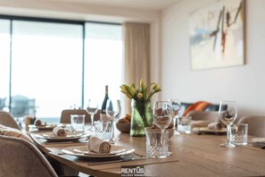 Une table à manger dressée pour un repas, avec des assiettes, des verres et une bouteille, dans une pièce dotée d’une grande fenêtre et d’un tableau au mur.