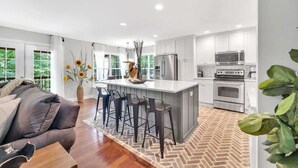 Una cocina moderna con armarios blancos, una isla central con taburetes de bar, electrodomésticos de acero inoxidable y un suelo con estampado de espiga.