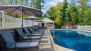 Un espace aménagé au bord de la piscine, avec des chaises longues et des parasols, entouré d’une clôture en bois peinte en blanc et d’arbres.