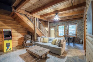 L’intérieur d’une cabane en rondins, avec un escalier en bois, un canapé, une table basse et une borne d’arcade Pac-Man.