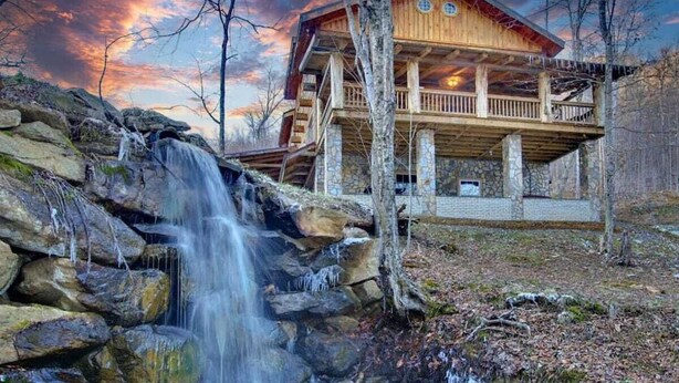 Una cabaña de madera con una cascada frente a ella, rodeada de árboles.