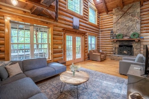 A log cabin interior with a fireplace, a sofa, a round coffee table, and large windows.