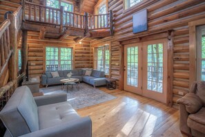 Interior de una cabaña de madera con sala de estar, un balcón y ventanas amplias.