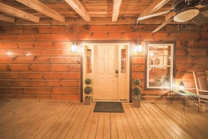 A log cabin interior with a wooden door, two wall-mounted lanterns, a small table with a lantern, and a chair.