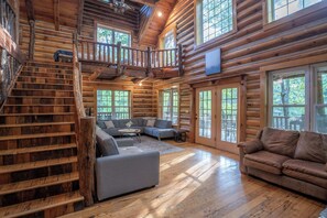 L’intérieur d’une cabane en rondins comprend un escalier, un salon avec un canapé et une table basse, ainsi que de grandes fenêtres.